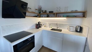 Een compacte keuken met witte kasten, een zwart inductiekookveld, een inbouwoven, een spoelbak en een aanrecht met diverse keukenbenodigdheden.