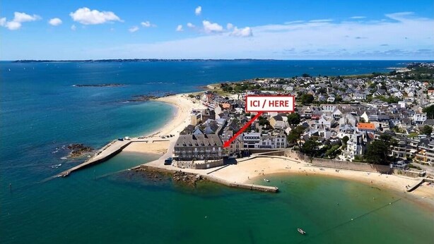 Luchtbeeld van een kustplaatsje met een strand, een pier en een gebouw met de tekst 'ICI / HERE'.