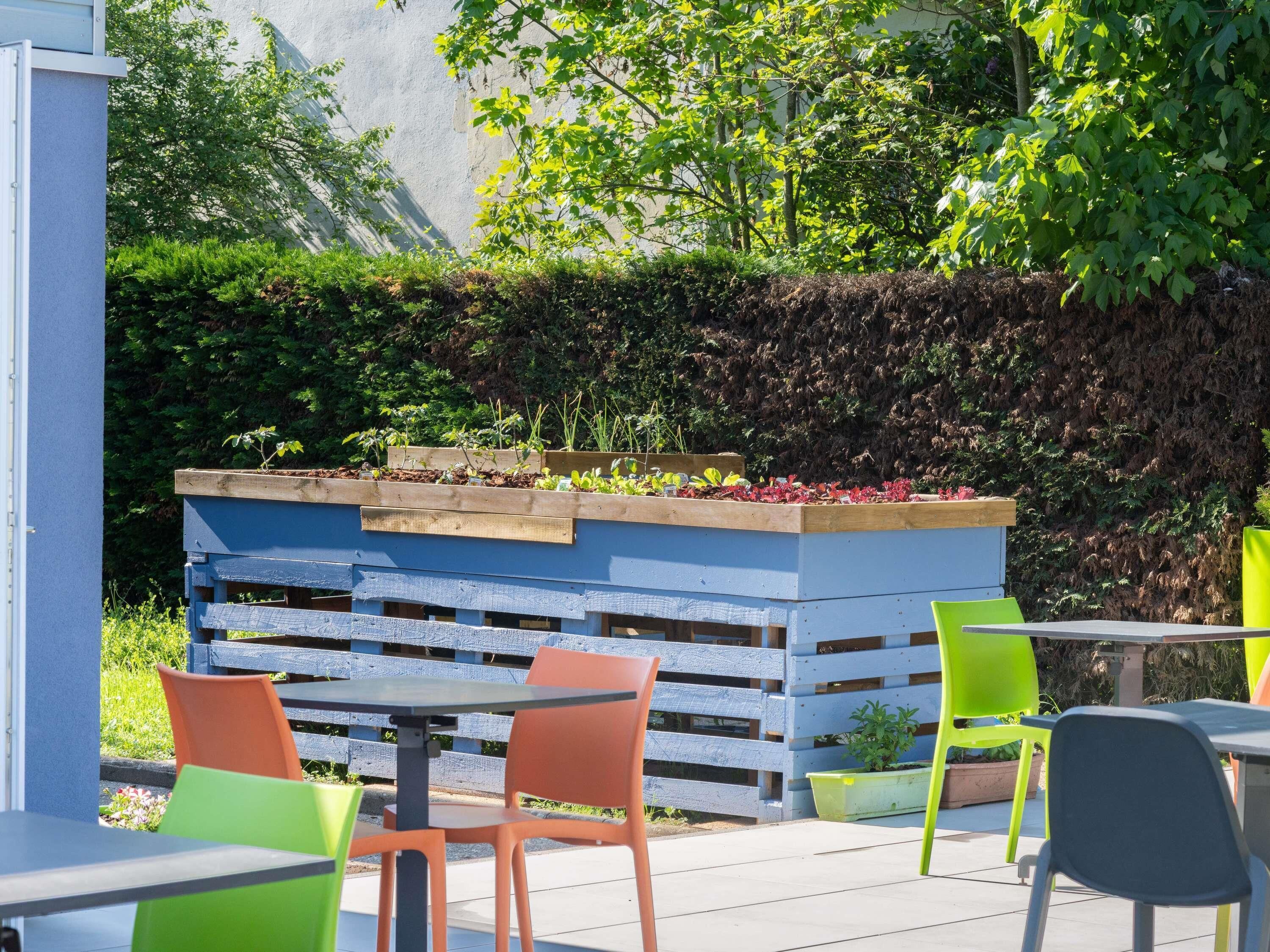 An outdoor seating area with wooden tables and chairs, a raised planter with various plants, and a blue wall in the background.