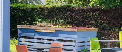 An outdoor seating area with wooden tables and chairs, a raised planter with various plants, and a blue wall in the background.