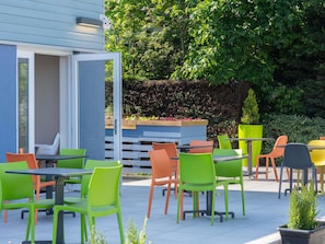 An outdoor patio with multiple tables and chairs, a wooden planter with plants, and a hedge in the background. - Ibis Budget Sélestat (Sélestat)