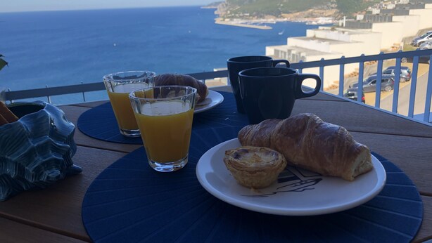 Une table avec un croissant, deux verres de jus d’orange et une tasse noire, donnant sur une vue sur la côte.