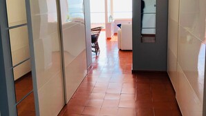 Un couloir au sol carrelé, avec un ventilateur de plafond, et une vue sur une pièce où se trouvent un lit et une chaise.