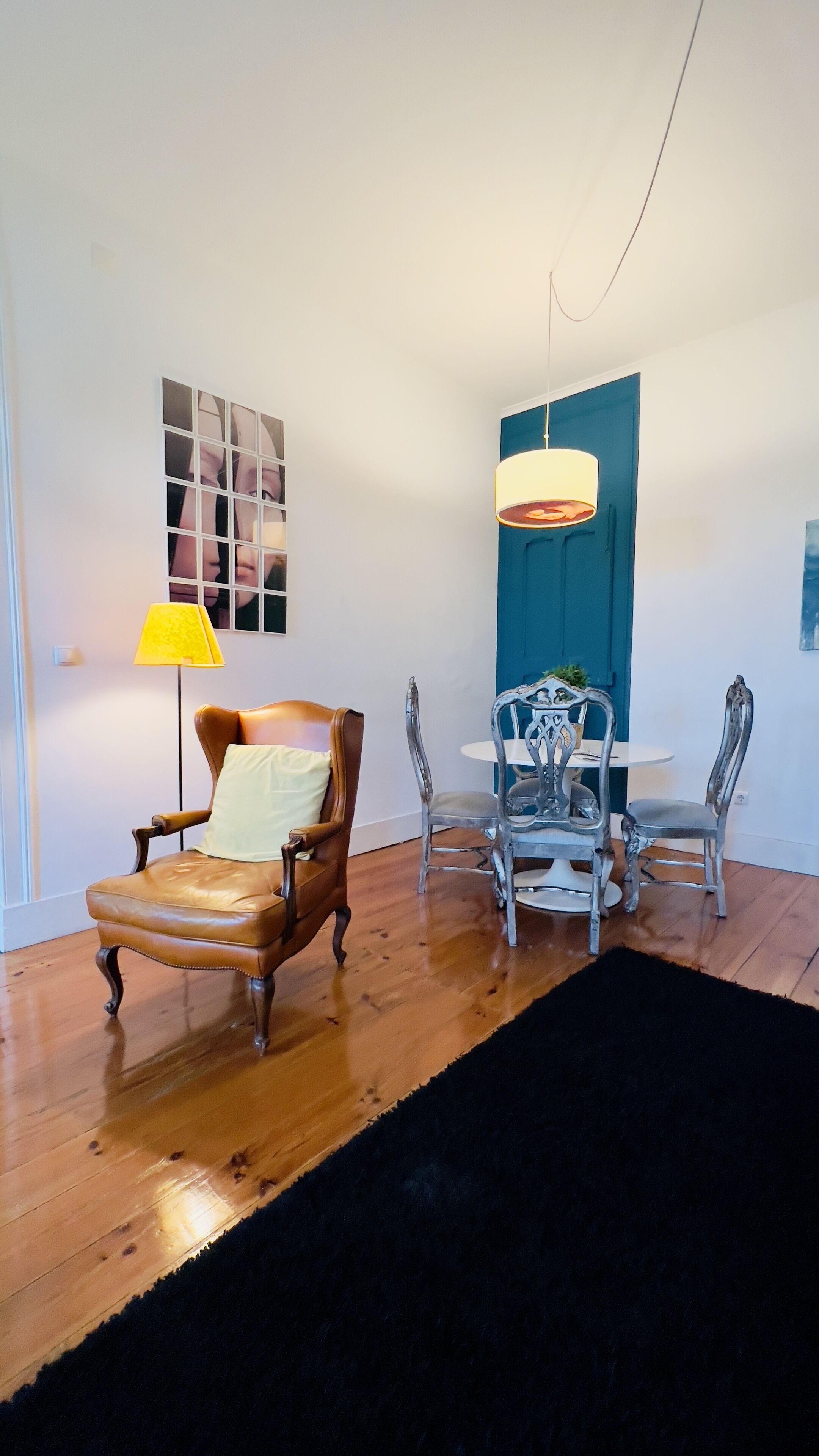 Una habitación con suelo de madera, un sillón de cuero marrón, una mesa redonda con sillas, una puerta azul y una lámpara de pared.