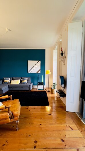 Een woonkamer met een donkerblauwe accentmuur, een grijze bank, een houten vloer en een gele vloerlamp.