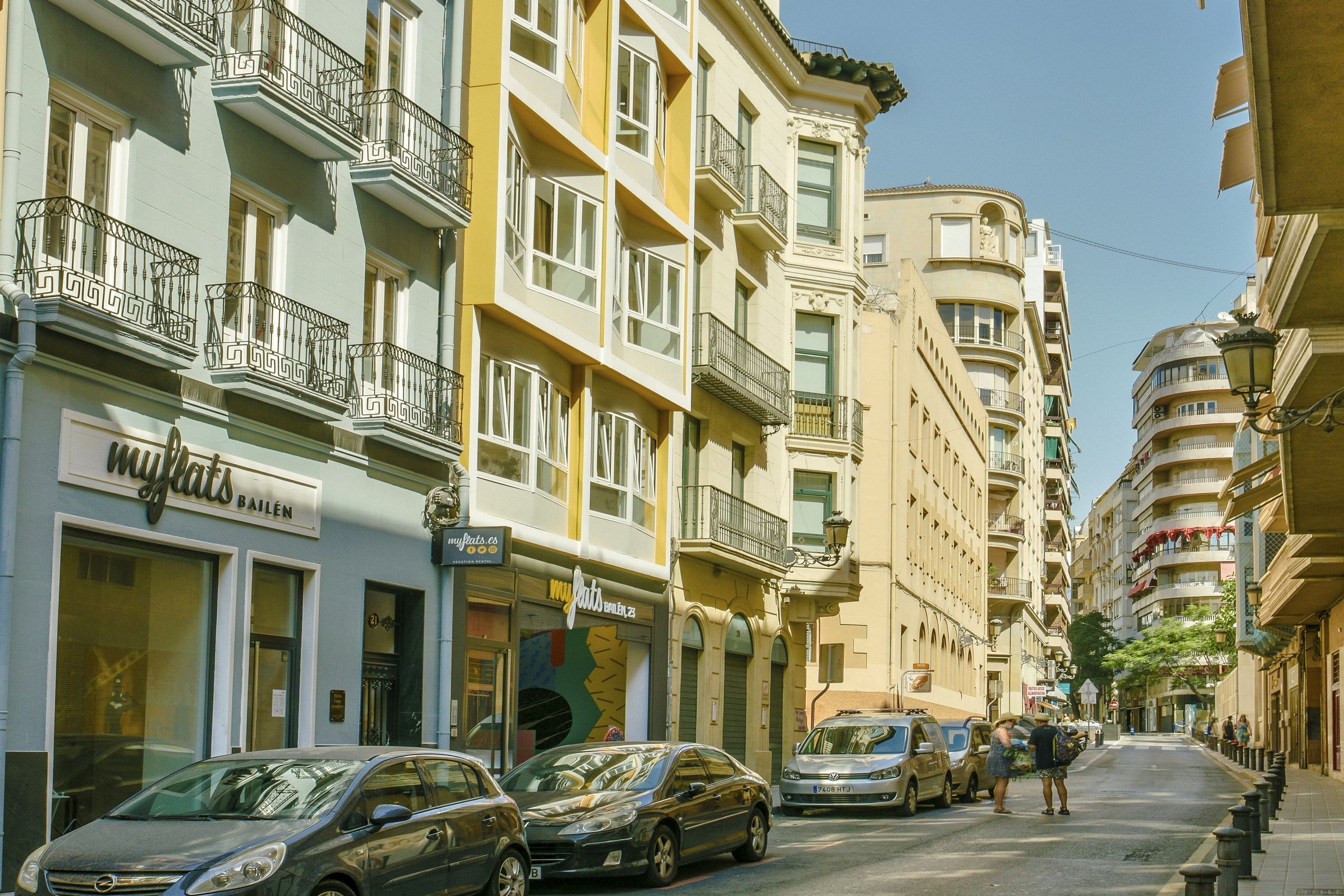 Une rue bordée d’immeubles à plusieurs étages, avec des voitures garées et un ciel dégagé.
