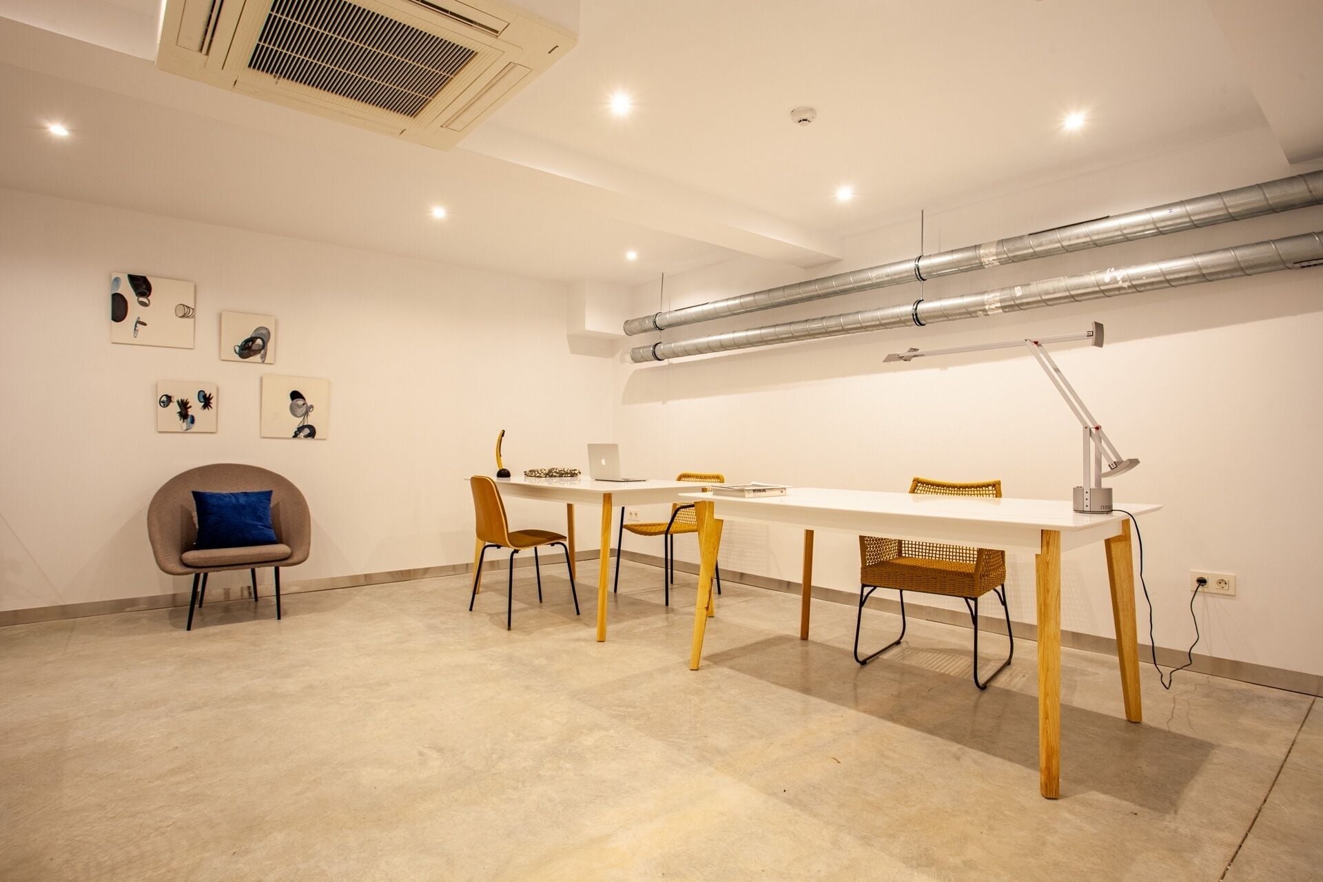 Un espacio de oficina moderno con un escritorio blanco, sillas y una computadora portátil. Se observan rejillas de ventilación en el techo y una lámpara fijada a la pared.