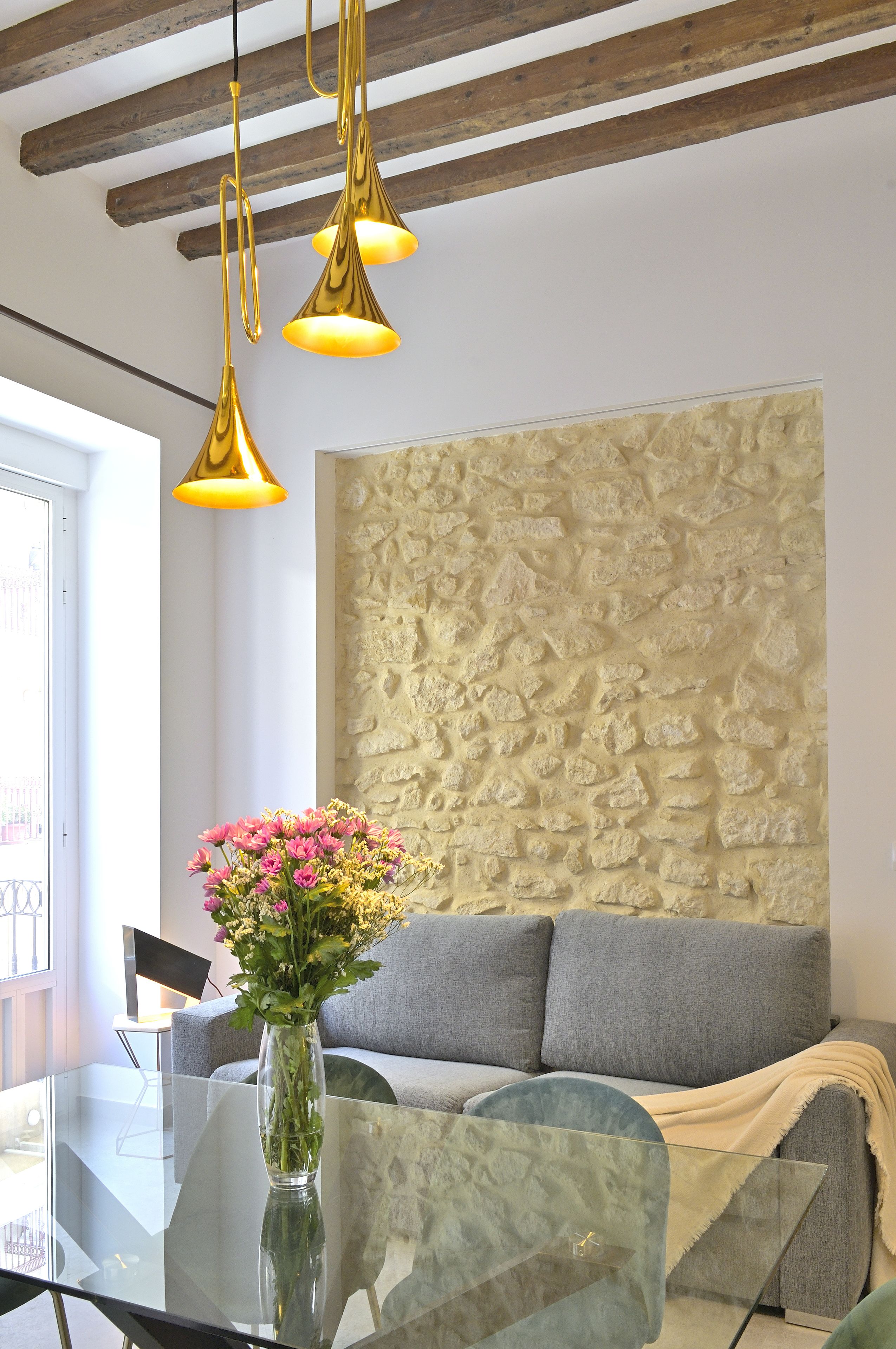 A modern living room with a stone accent wall, a glass coffee table with a vase of flowers, and a grey sofa.