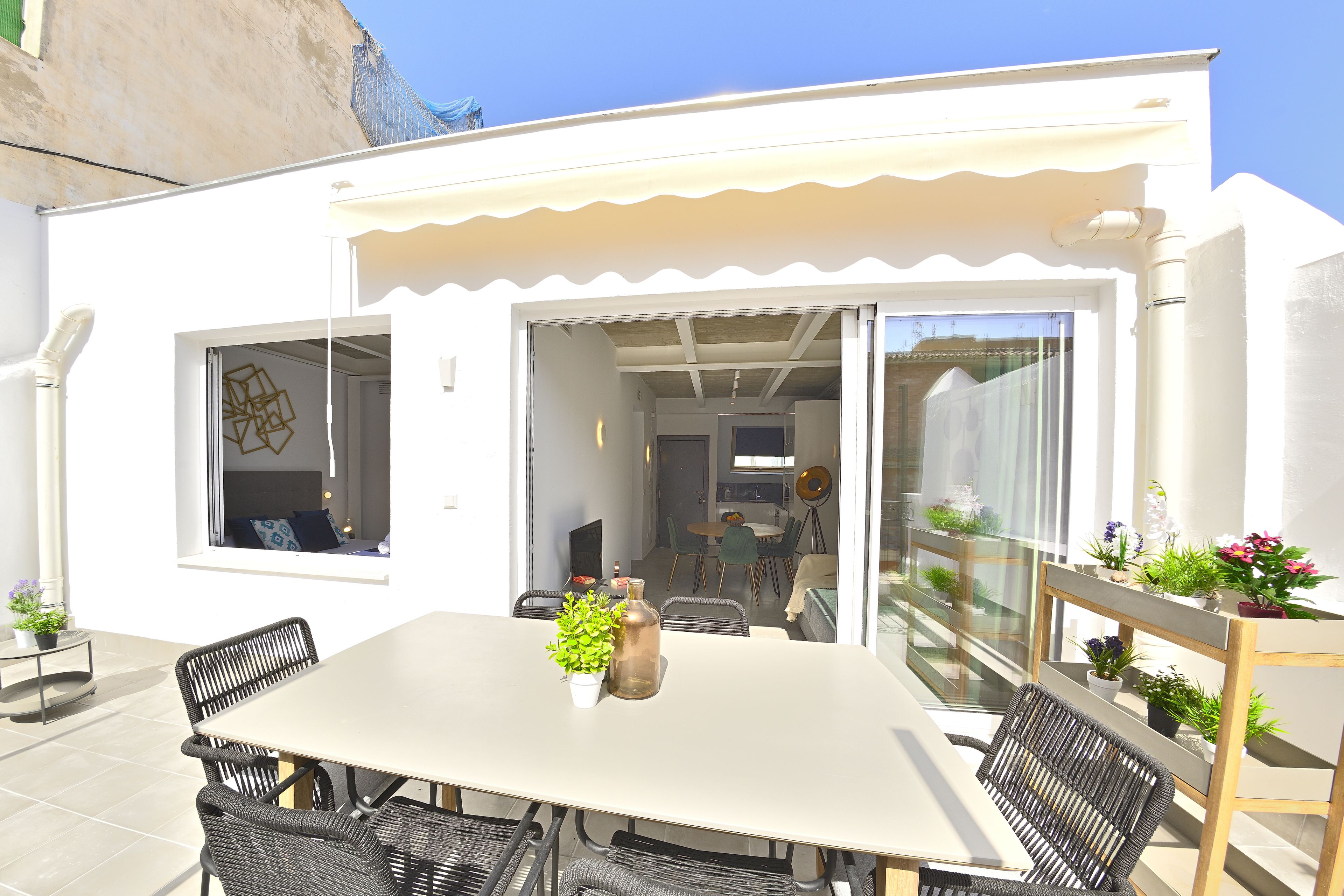 A modern outdoor dining area with a white table and black wicker chairs, surrounded by potted plants and a glass railing.