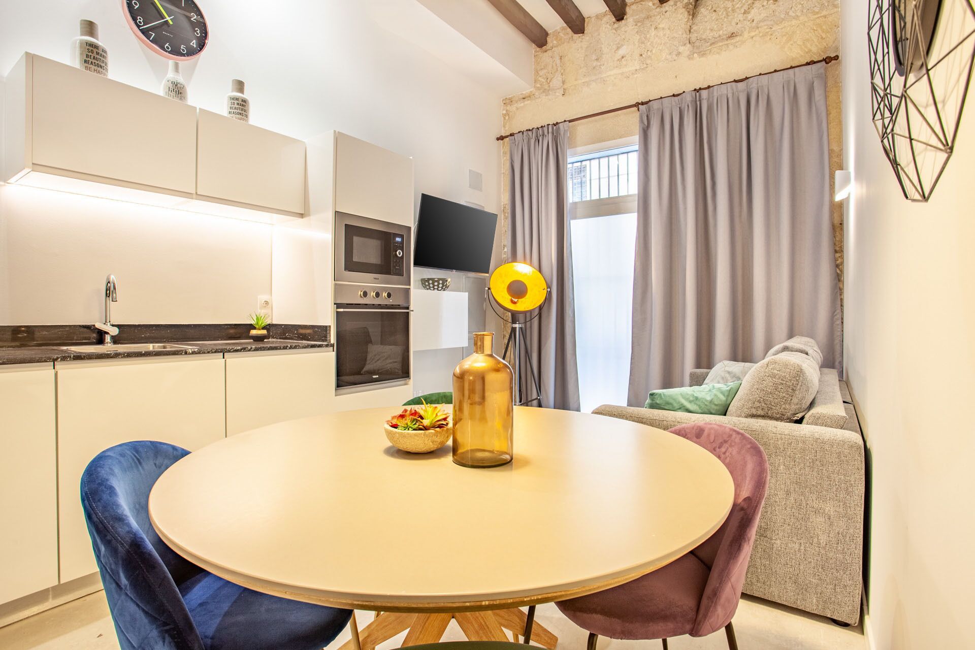 A modern kitchen with a built-in oven and microwave, a dining table with chairs, and a living area with a sofa and a TV.
