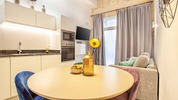 A modern kitchen with a built-in oven and microwave, a dining table with chairs, and a living area with a sofa and a TV.