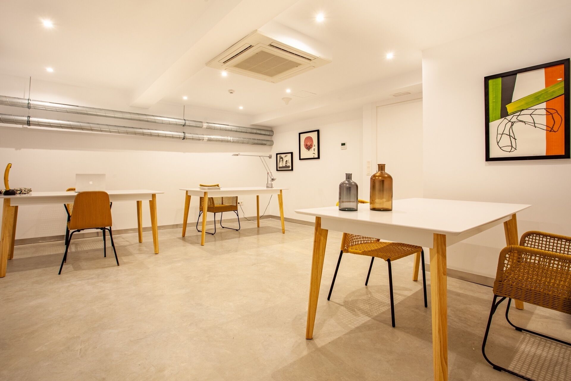 A modern, well-lit meeting room with white tables and chairs, a visible air conditioning unit, and artwork on the walls.