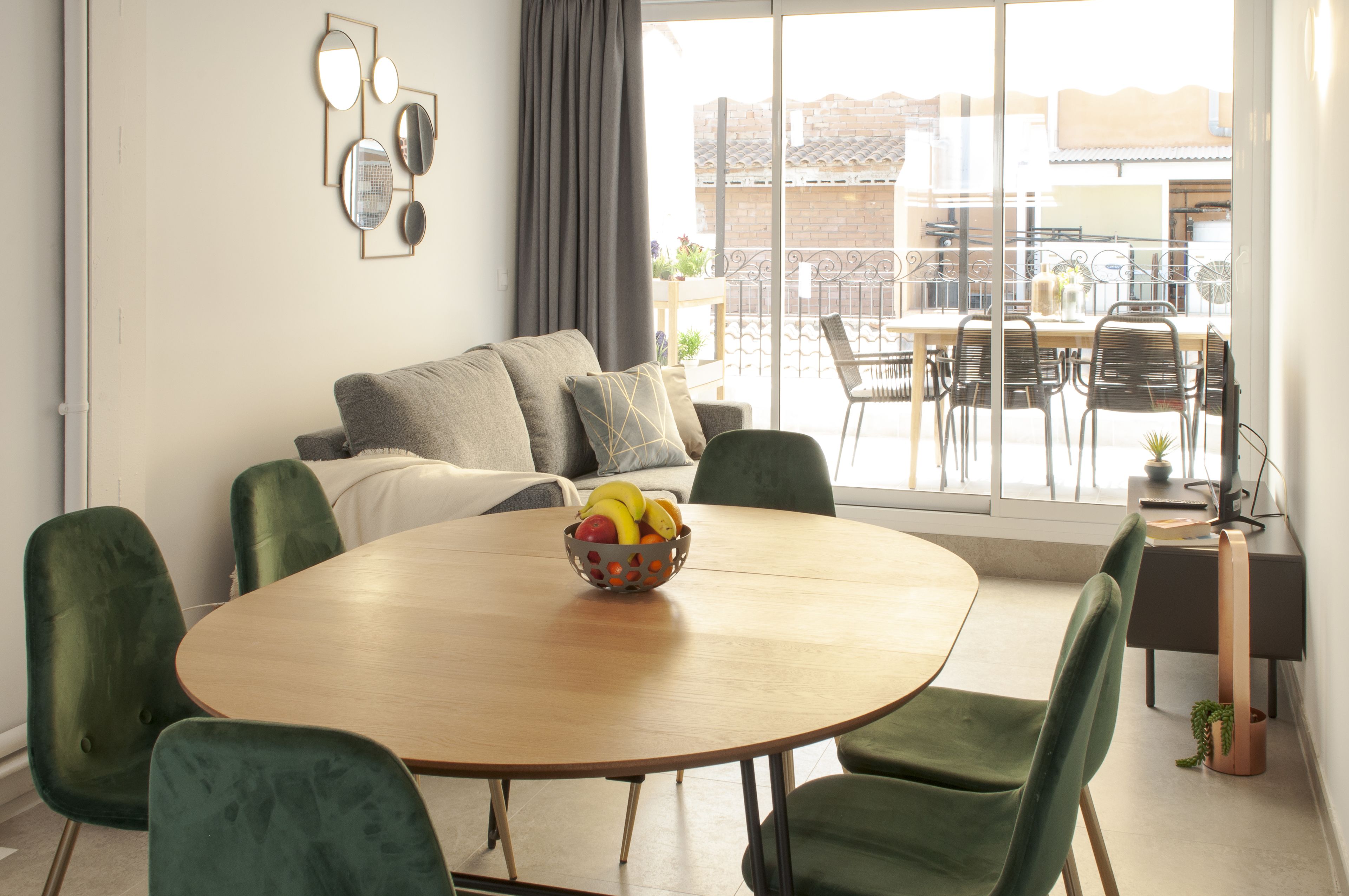 A dining area with a round table, green upholstered chairs, and a bowl of fruit.