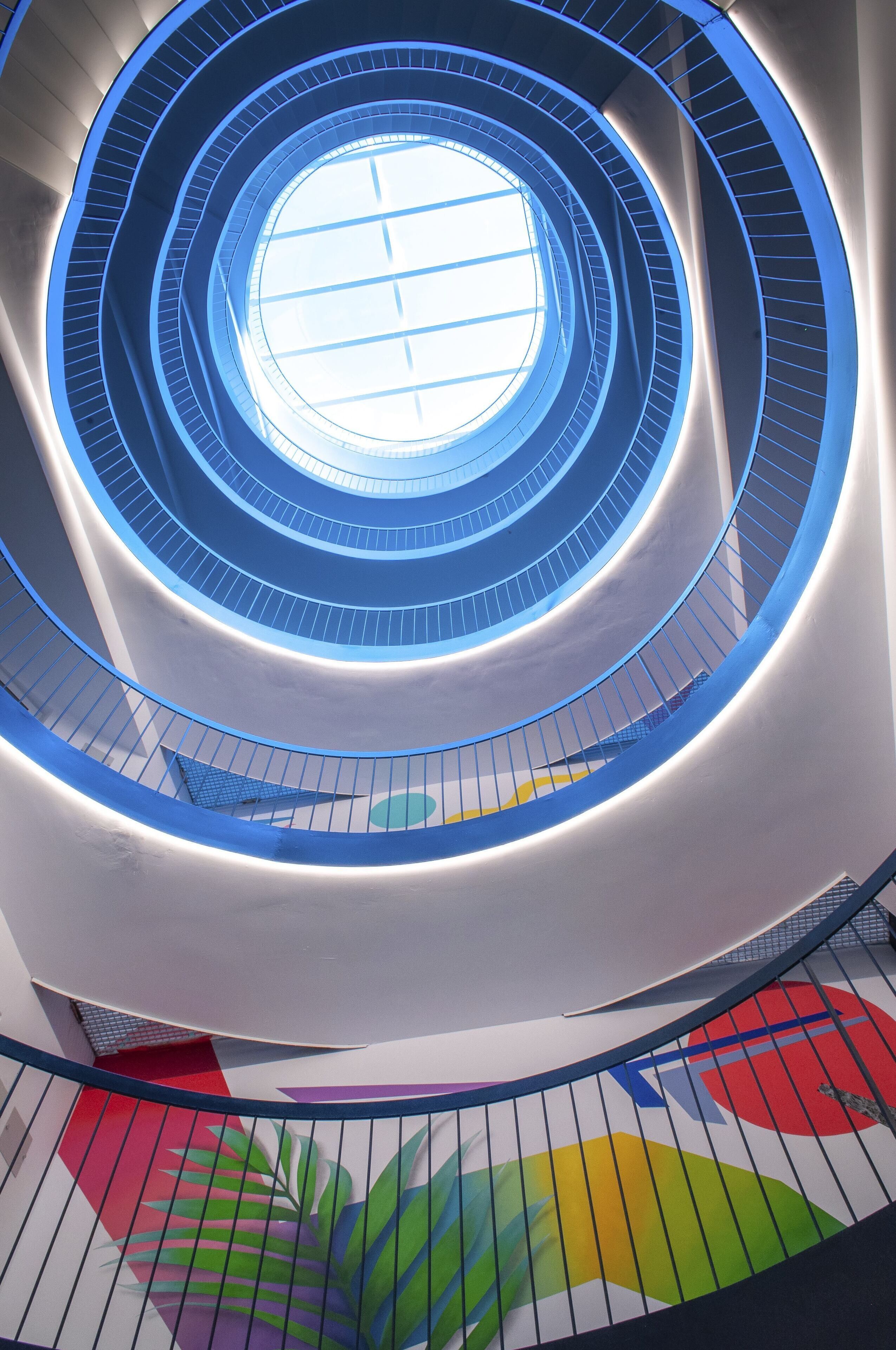 A spiral staircase with a central skylight, surrounded by a white wall with a colorful mural.