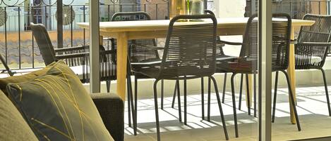 A balcony with a wooden table and black metal chairs, a sofa with cushions, and a view of buildings in the background.