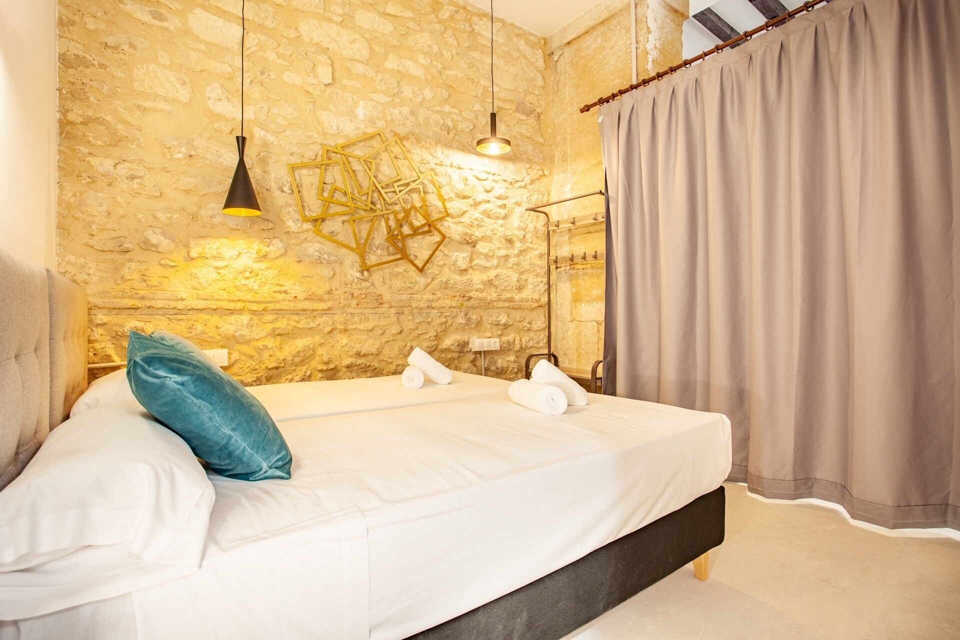 A bedroom with a stone wall, a bed with white linens, a blue pillow, and a hanging light fixture.