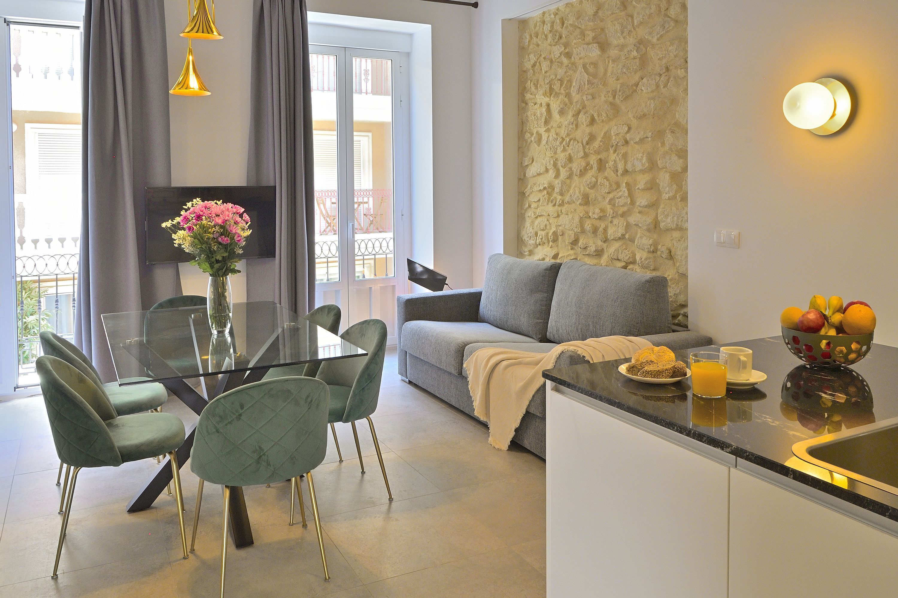 A modern living room with a grey sofa, a glass dining table with green chairs, a TV mounted on the wall, and a stone textured wall.