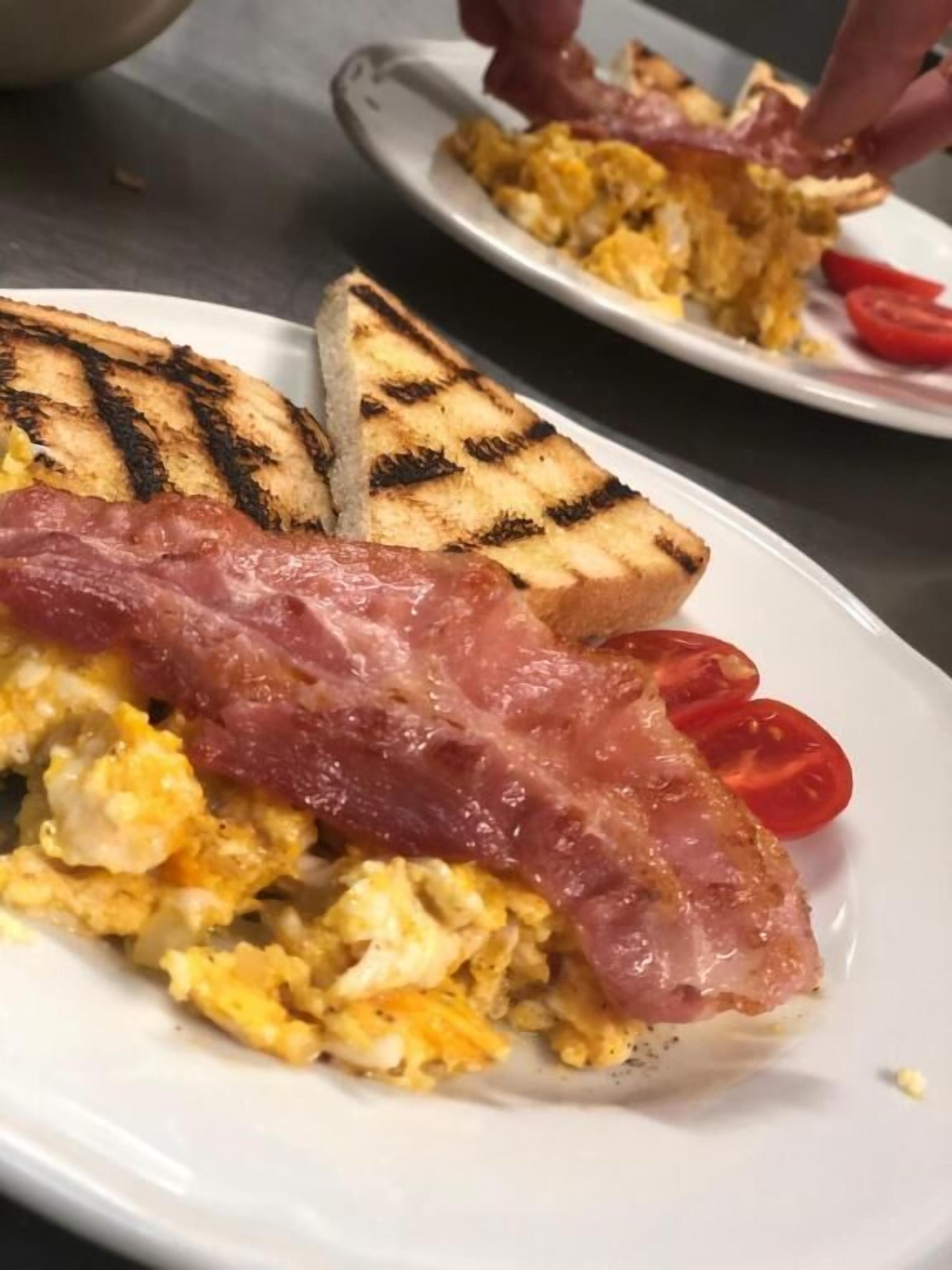 A breakfast plate with scrambled eggs, bacon, grilled toast, and tomato slices.