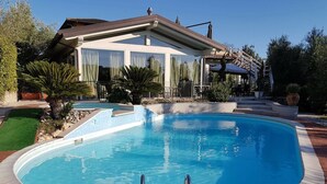 Een zwembad met helderblauw water, omgeven door een terras van baksteen, en een huis met een overdekte patio op de achtergrond.