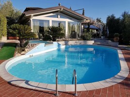 Una piscina con agua azul cristalina, rodeada por un suelo de ladrillo, y una casa con un porche cubierto al fondo.