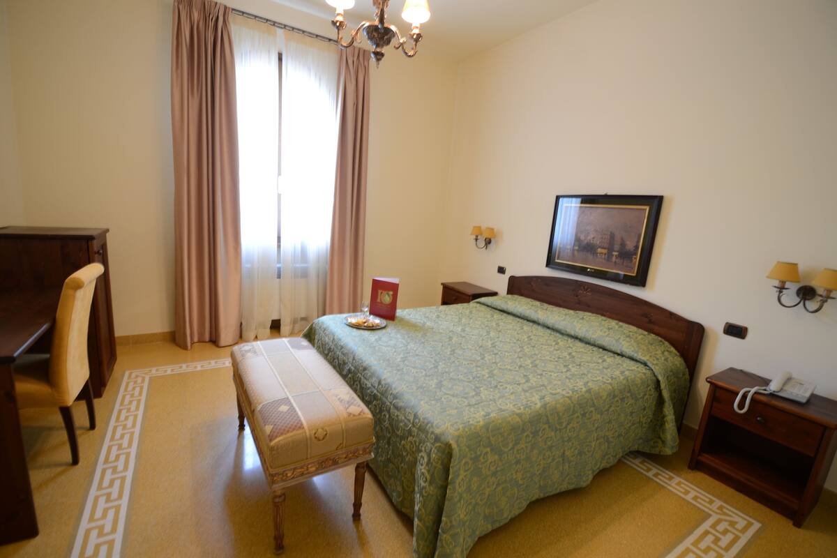 Habitación de hotel con cama, escritorio, silla, mesita de noche con lámpara y un cuadro en la pared.
