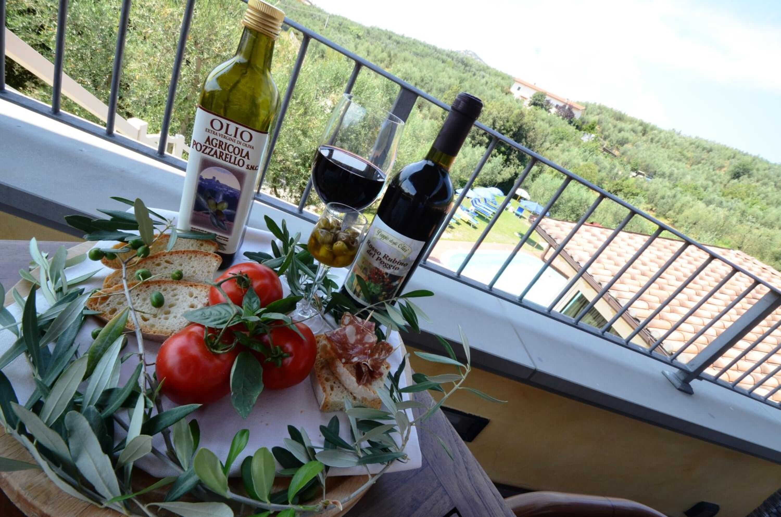 Ein Tisch mit Olivenöl, einer Weinflasche, einem Weinglas, Brot, Tomaten und Oliven auf einem Balkon mit Blick auf eine Hügelkette.