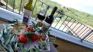 Ein Tisch mit Olivenöl, einer Weinflasche, einem Weinglas, Brot, Tomaten und Oliven auf einem Balkon mit Blick auf eine Hügelkette.