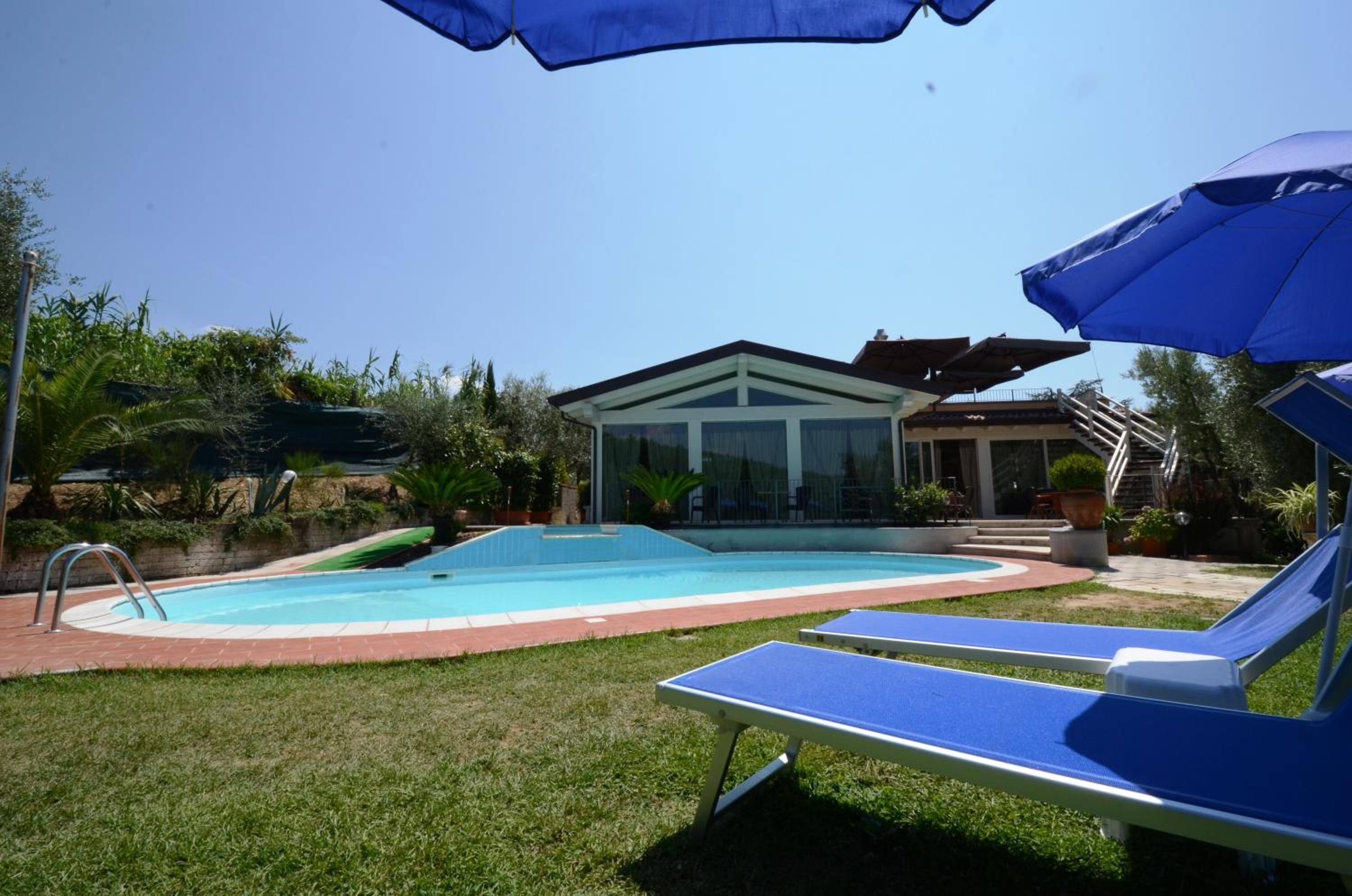 Une piscine entourée de chaises longues et de parasols, elle-même bordée de verdure et d’un bâtiment doté d’une structure en verre.