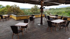 Eine Terrasse im Freien mit Holztischen und -stühlen, einem Metallgeländer und Blick auf Grünflächen und Hügel.