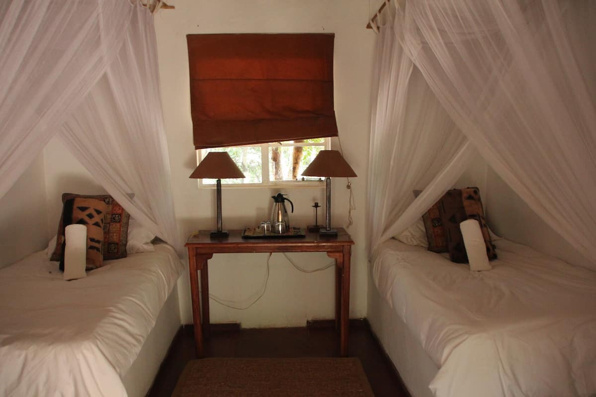 A room with two beds, a window with a brown blind, and a table with a lamp and a coffee pot.