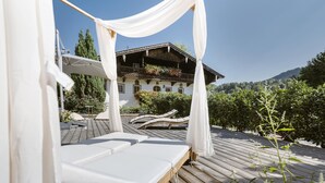 A wooden deck with white cushioned lounge chairs, a white canopy, and a building with balconies in the background. - Bauernhaus-Schloss Wagrain (Ebbs)