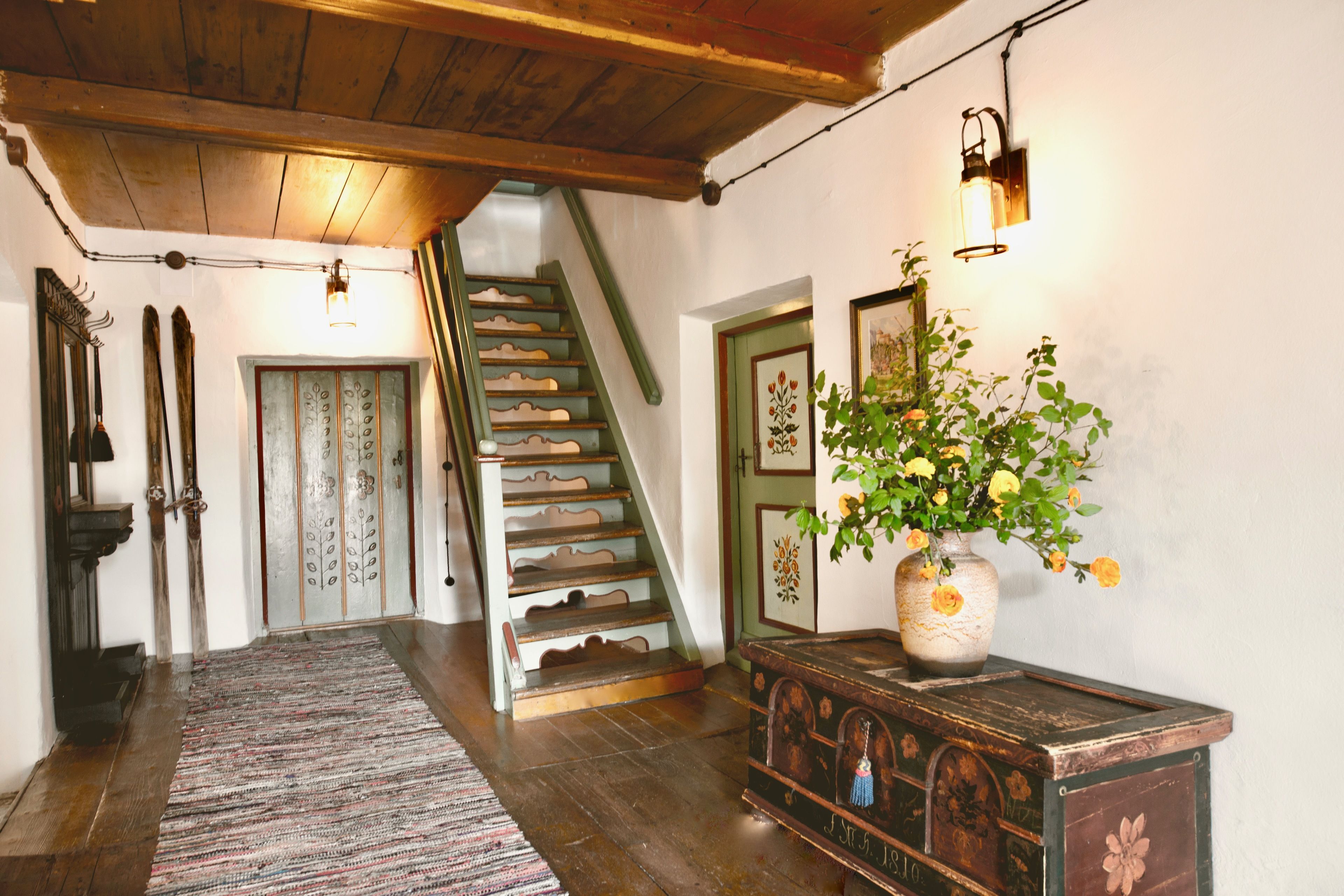 A wooden staircase with a green railing leading to a door with floral decorations. A patterned rug is on the floor, and a vase with yellow flowers is on a wooden cabinet.