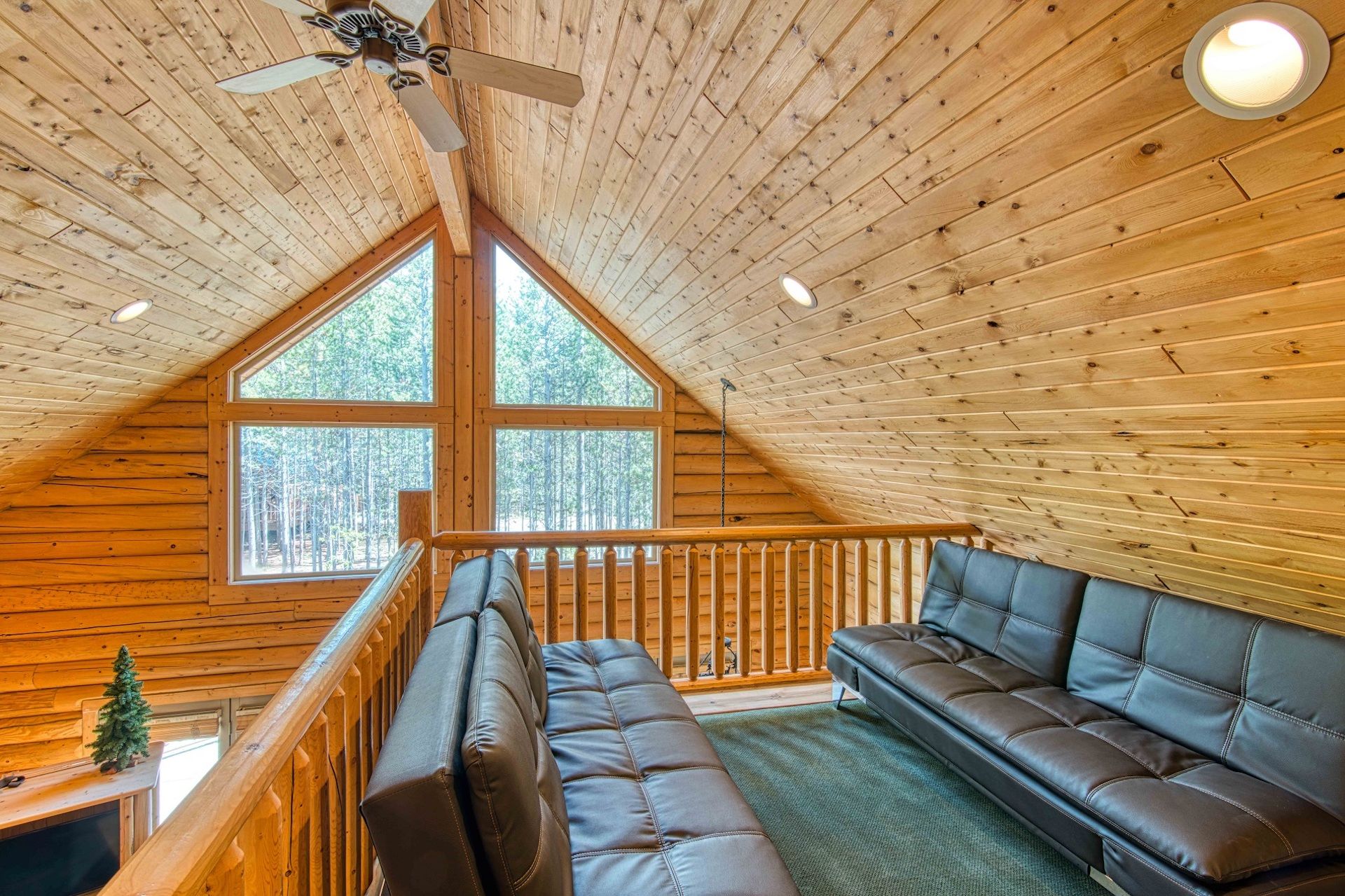 A wooden cabin interior with a loft bedroom, a ceiling fan, a large window, and a sofa.