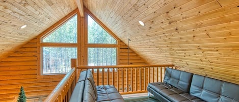 A wooden cabin interior with a loft bedroom, a ceiling fan, a large window, and a sofa.