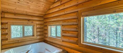 Interior de una cabaña de troncos con una bañera grande, dos ventanas y vista a los árboles.