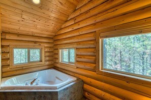 A log cabin interior with a large bathtub, two windows, and a view of trees.