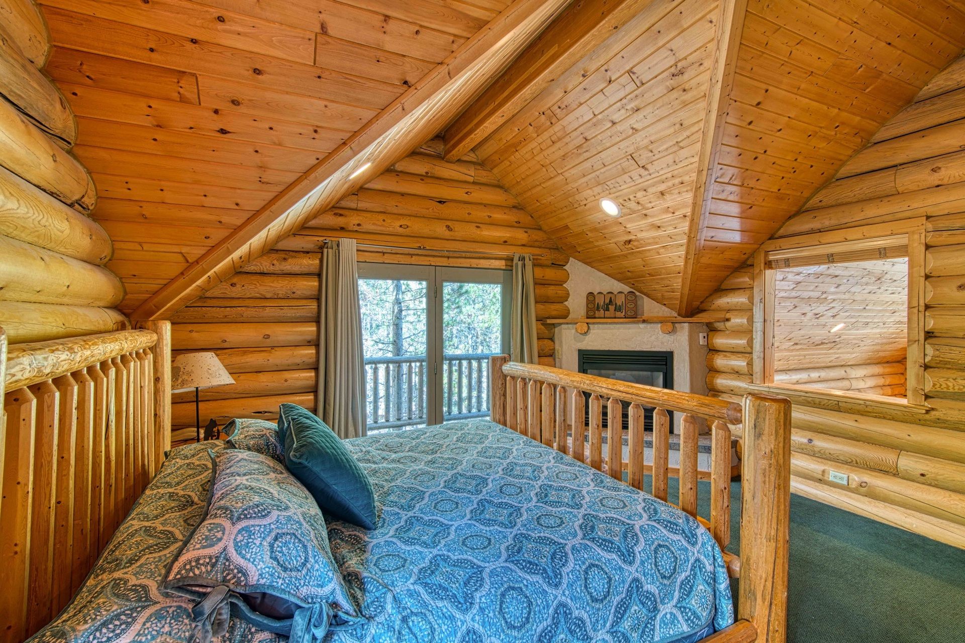 A log cabin bedroom with a bed, a fireplace, and a window overlooking a deck.