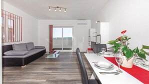 Uma sala de estar moderna com um sofá, uma mesa de centro de vidro e uma área de jantar com mesa posta para dois.
