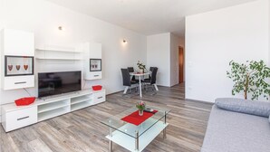 Uma sala de estar moderna com uma televisão de tela plana, uma mesa de centro de vidro, um sofá cinza e um espaço de jantar com mesa e cadeiras.