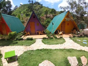 Tres pequeñas cabañas triangulares con techos verdes, dispuestas en fila con un sendero arenoso entre ellas.