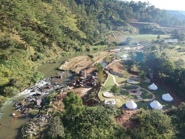 Un campamento a orillas del río, con tiendas de campaña y un pequeño edificio, rodeado de un bosque denso y campos.