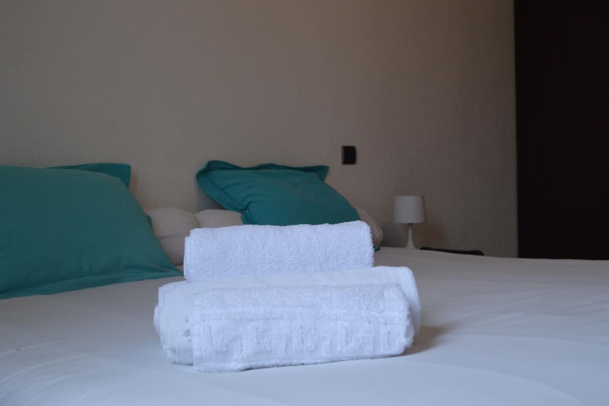 Un lit bien fait, agrémenté d’oreillers blancs et turquoise, d’une serviette blanche et d’une petite table de chevet avec une lampe.