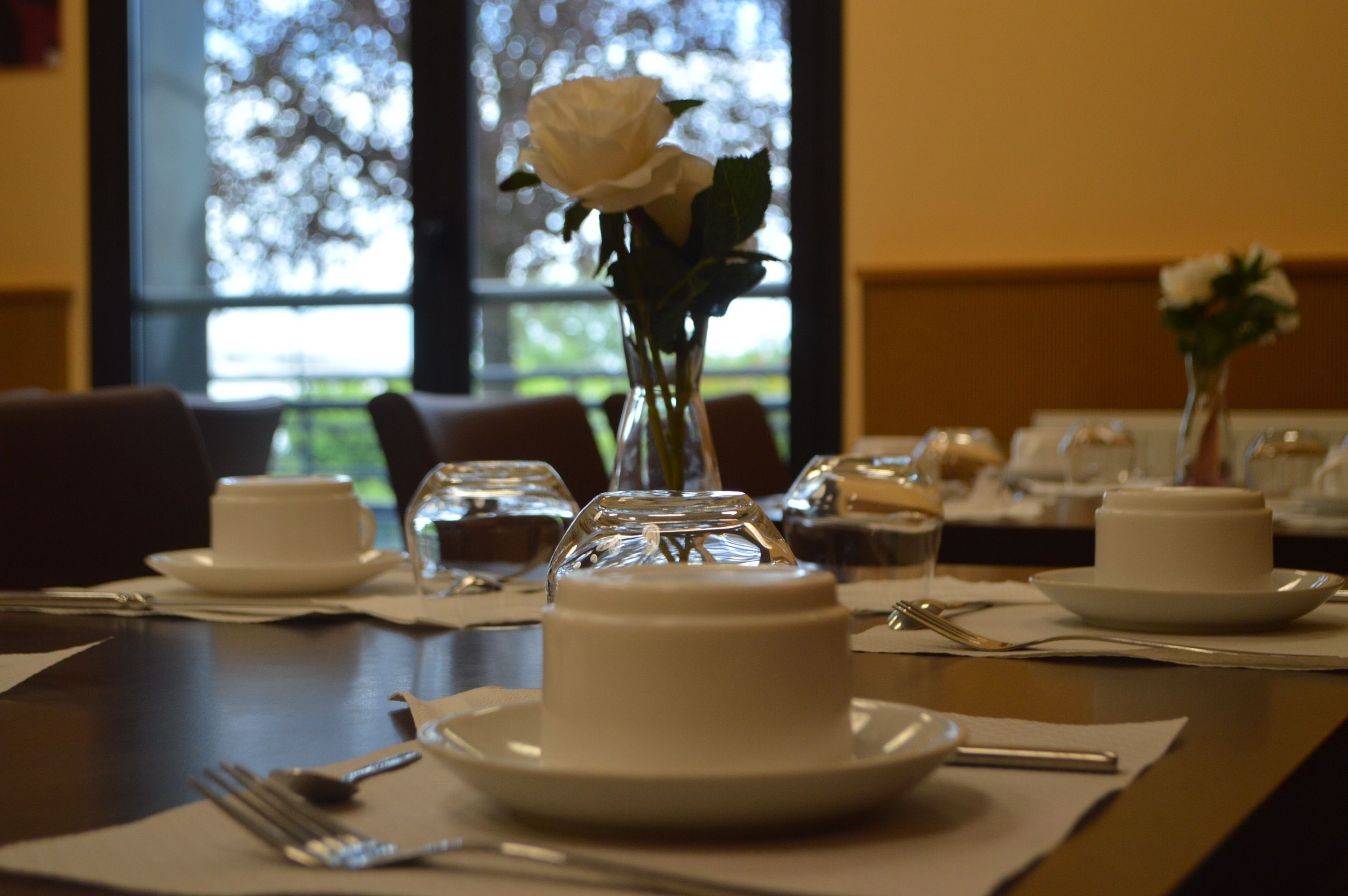 Una mesa de comedor dispuesta con platos blancos, tazas y un jarrón con una rosa blanca.
