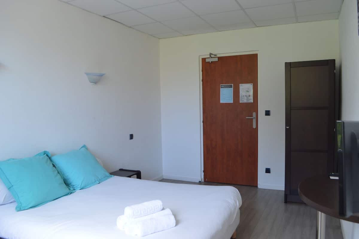Une chambre d’hôtel avec un lit, deux oreillers, une table de chevet, une porte en bois et un téléviseur posé sur un meuble.