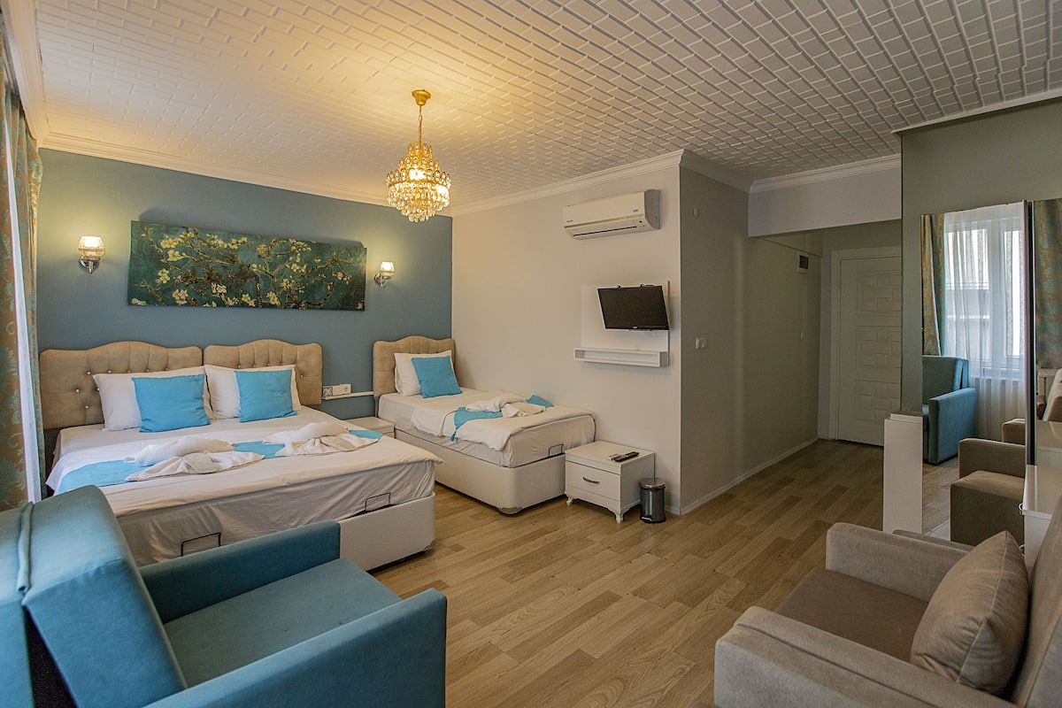 A modern hotel room with a large bed, two smaller beds, a seating area with sofas, a chandelier, and a television.