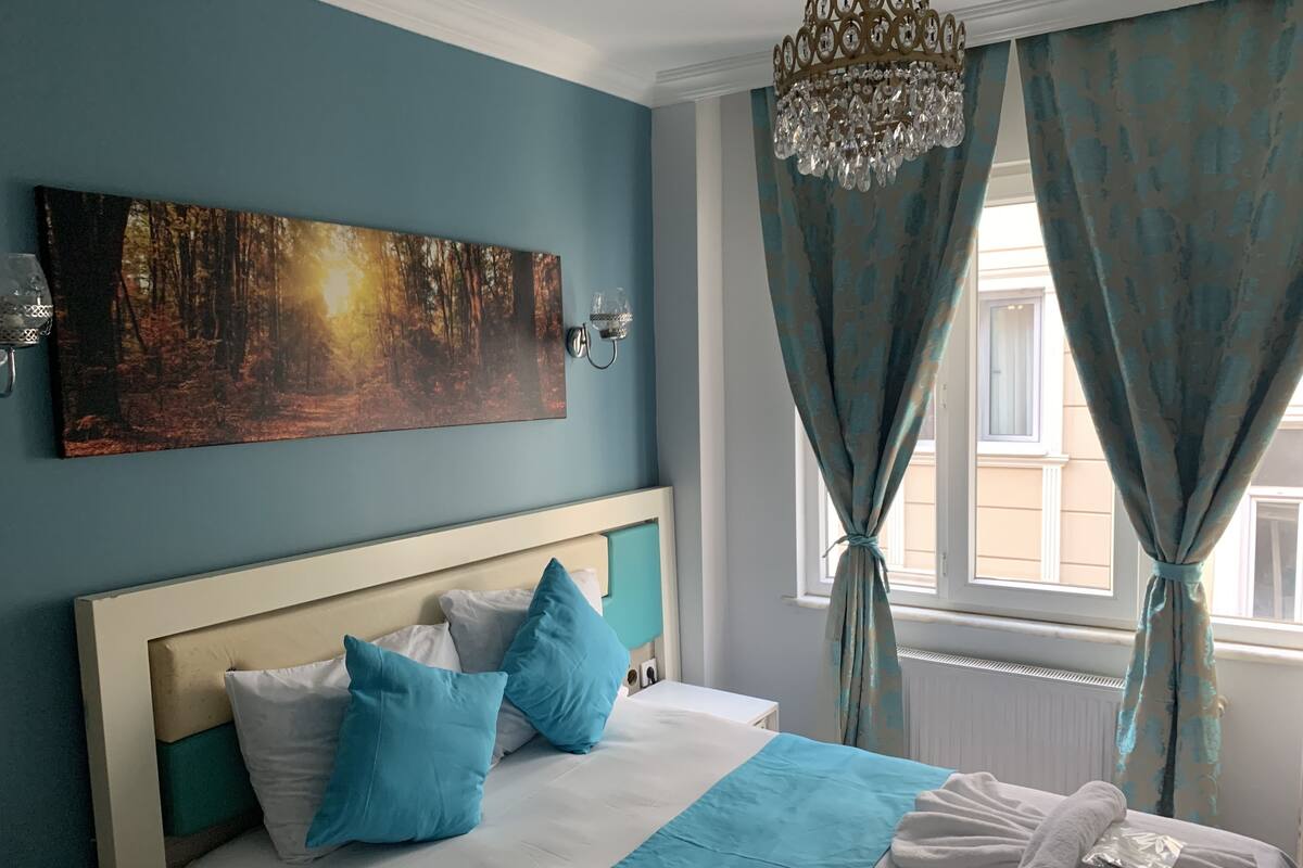 A bedroom with a bed, pillows, a painting, a chandelier, and curtains.