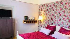 Une chambre à coucher avec un lit, un téléviseur posé sur un meuble, une étagère avec des objets décoratifs et un papier peint à motifs floraux.