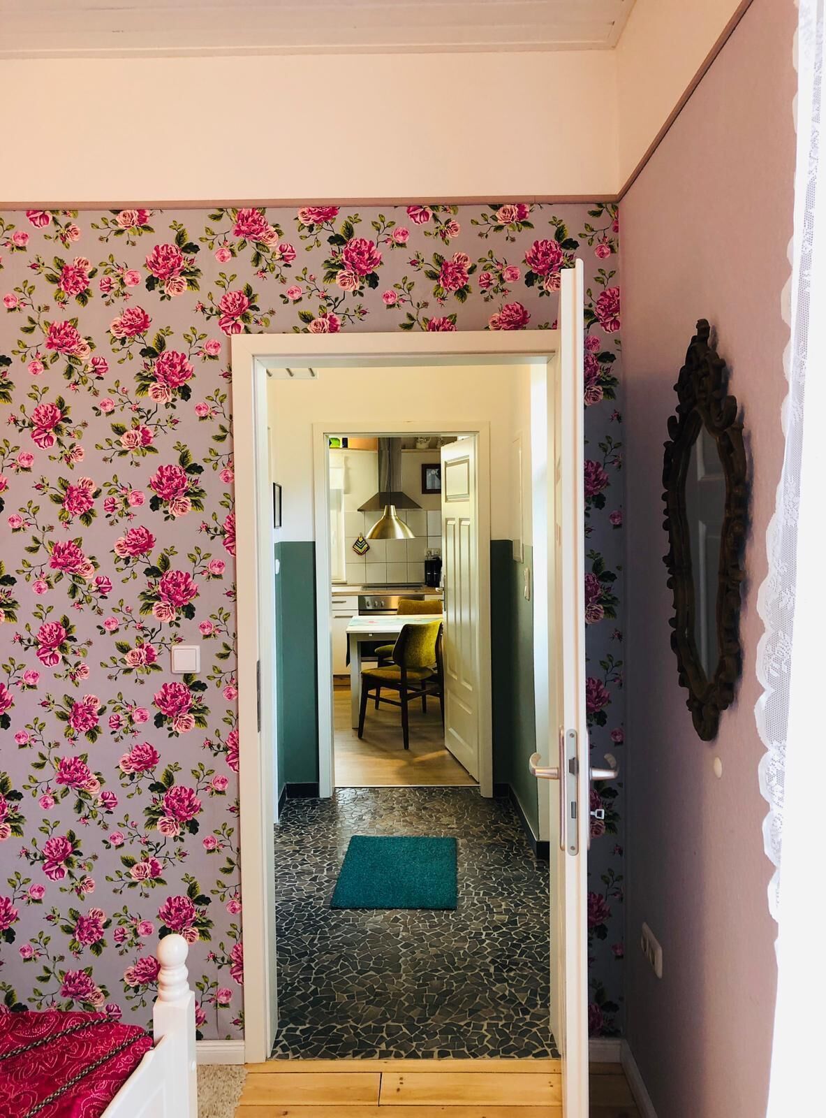 Ein Zimmer mit floraler Tapete, einem hölzernen Bett, einem grünen Teppich und Blick in einen Küchenbereich mit Esstisch und Stühlen.