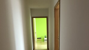 Un pasillo con suelo de madera y paredes blancas que conduce a una habitación con una pared verde y una pequeña mesa con una lámpara.