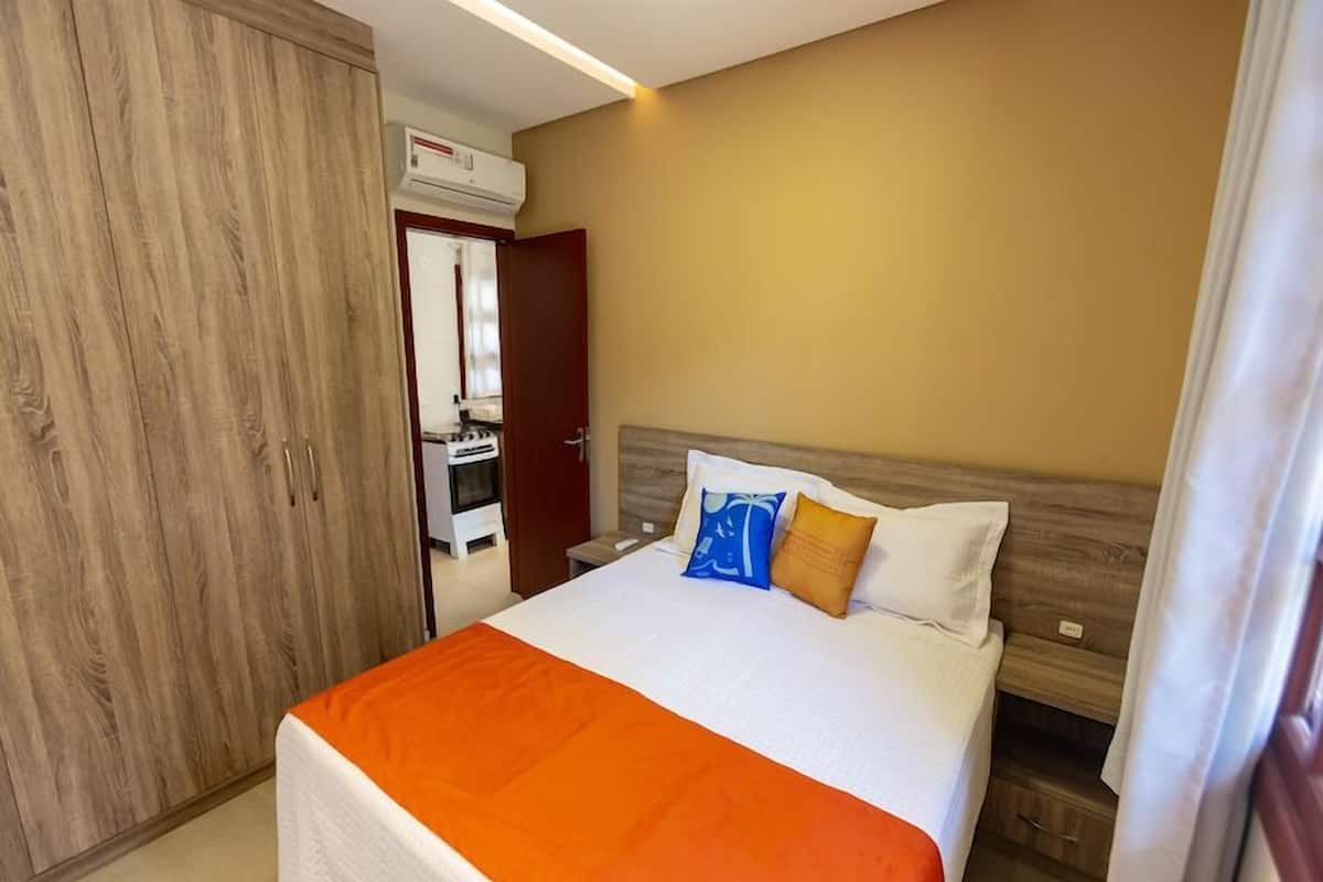Une chambre d’hôtel avec un lit, une couvre-lit orange, une armoire en bois et, au fond, une petite cuisine.